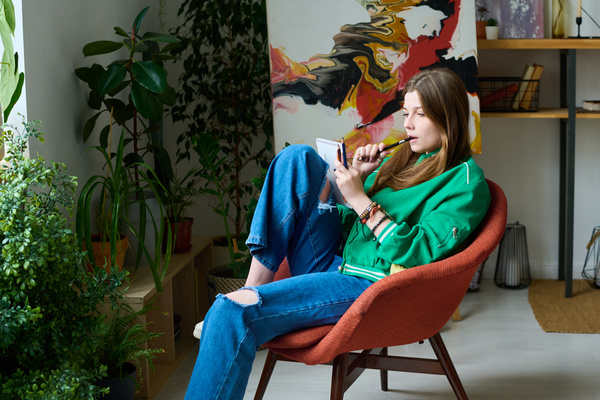 Zoomer girl with brown hair in a green jacket and jeans with holes is sitting on a red chair with one foot on the seat and thoughtfully looks at her drawing with a pencil in her mouth